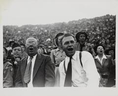 Image of a large crowd. There are two men clear in the foreground. The man on the left wears a striped suit jacket and tie. The man on the right wears suspenders and a long dark tie.&nbsp;