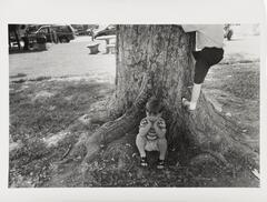 A black and white image of a boy sitting at the base of a tree. Another child, with only the lower half of his body visible, is climbing the tree and a table and park benches are partially visible behind the tree. 