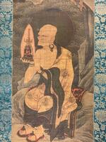Long hanging scroll with printed gold and blue cloth on either side of the image. Image depicts an aged man sitting on a rock holding a relic in his right hand. A halo sits around his head. Above and behind him are waves and more rocks, suggesting he is seated on a rocky outcropping near a mass of water. The colors are heavily faded, though the robe around the man is primarily gold, with green and red accenting.