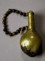 Small brass container with a round body and a short, narrow neck. The wooden stopper is attached to the container by a string of brass beads. The body of the container is decorated with a design of incised concentric circles. 