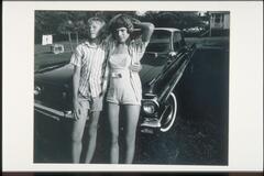 A photograph of two adolescents, one male, one female, standing side-by-side in front of an automobile.