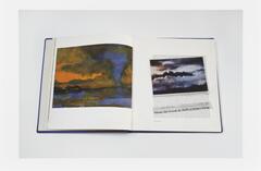 Image of a book lying open to a page featuring painted clouds. A newsclipping featuring a photo of clouds lies on the book across the open page.  