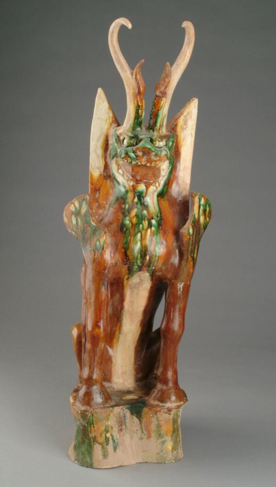 An earthenware figure of a zoomorphic form consisting of a lion-like body with strong hoofed feet, wings, horns, and a dragon-like face bearing teeth in its open mouth. It is seated on a rock-like base, and covered in amber, green, and cream runny glazes. One of a pair with 2004/2.132.2.