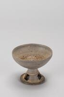A ceramic rounded bowl with a tall, trapered cylindrical stand with a wide lip. The base is detailed with staggered square cut-outs that run from the lip to the middle of the base.<br />
<br />
This is a dark-gray, single-tiered, perforated, high-fired stoneware stem cup. The body of the cup curves upwards, and the rim has a sharp edge, while the inner surface of the mouth is tapered. A deep incised line divides the pedestal into its upper and lower halves; the lower half features square perforations. A natural glaze is pooled at the base of the cup, while the bottom of the outer surface shows traces of rotation and water smoothing.
<p>[Korean Collection, University of Michigan Museum of Art (2017) p. 65]</p>
