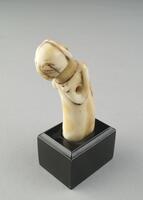 One of a pair of delicately carved, hippopotamus ivory statuette-pendants.  This female figure's head and upper body lean slightly forward, as opposed to her counterpart who stands upright.  Both, however, have been carved by the same hand and display the hallmark characteristics of the northeastern Luba stylistic form, namely: the round head with a convex face, large, coffeebean-shaped eyes, a rectangular mouth with prominent lips, a cylindrical neck, and a coiffure decorated on the back with a cruciform pattern. Additionally, both female figures clutch their breasts in their hands, a pose commonly seen among Luba sculptures of women. The statuettes have been pierced through, allowing them to suspend from a string.