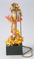 Carved wooden columnar figure wrapped with light colored wire. The top of the figure is decorated with leather strands of yellow and orange beads. The limbs of the figure are formed by strands of white and yellow seed beads terminating in large yellow and orange beads. There is a large string attached to the figure, perhaps to wear around a person's neck. 