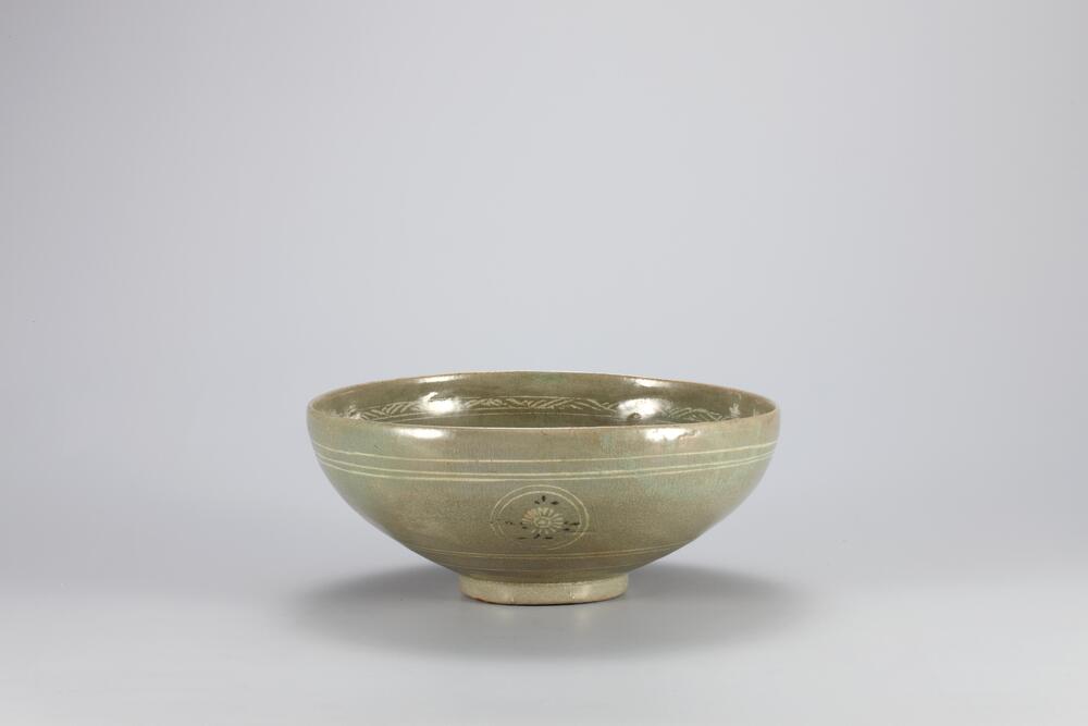 <p>This bowl is a fine example which displays the excellent decorative techniques applied to Goryeo celadon by its magnificent decorations: a band of foliage design right below the mouth, the four pomegranate designs on the inner wall, and a chrysanthemum floret on the inner bottom. On the outer surface, there are four sets of double concentric circles inlaid with white slip, each containing a chrysanthemum spray inlaid in black and white. The foot retains three quartzite spur marks. ere are cracks on the outer base due to the thickness of the wall. Glaze has been applied down to the foot and well-fused, while color is also evenly distributed.<br />
[<em>Korean Collection, University of Michigan Museum of Art </em>(2014) p.106]</p>
<br />
Shallow bowl with celadon glaze. Four concentric circles grace the exterior of the bowl, with a chrysanthemum roundel centered in the inner circle. Above these designs, three thinly incised bands stretch across the bowl parallel to the rim. In this location on the inside 