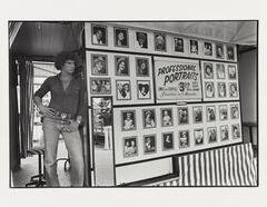 A black and white image of a man standing next to a wall covered in photographs with a sign advertising professional portraits.