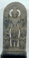 Vishnu stand in a strict unbending pose, samabhanga and has four hands.  Reading clockwise from the front right hand, he holds lotus, a club, a discus and a conch.  The lotus and conch are also personified with full standing figures at the base below his tow front hands, the lotus as a female figure to his right and the conch as a male figure to his left.  On the pointed arch behind the figure a flying figure holding garlands is carved in shallow relief to either side of his crown. He wears a diaphanous lower cloth, the folds of the garment are articulated with a flared section down the center.  He wears a long garland down to his knees, a sacred thread and various pieces of jewelry, including bracelets, armlets, a necklace and large earrings and an elaborate crown.<br />