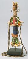 Columnar wooden figure decorated with beads. The figure has two small eyes and a mouth; the coiffure consists of a cluster of beads at the top of the head. There are small wire hoops on each side of the head. The neck is decorated with white seed beads while the torso is wrapped with red wire and blue beads. Strands of green and white beads form the limbs. The figure is attached to a large string of beads, possibly to wear the figure around a person's neck. 