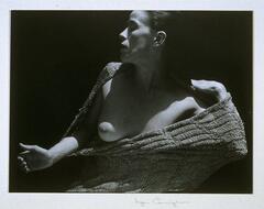This photograph depicts a woman standing in stark lighting and stretching knit fabric over her naked torso. 