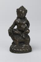 A miniature, cast bronze sculpture of Kubera, the god of wealth, seated sideways on a lion. Kubera sits in the lalitasana pose (the pose of royal ease, with one leg drawn up and the other relaxed); his right hand is outstretched to rest on the knee, while his left arm is akimbo and his hand rests on his hip. The base has a simple, single lotus petal design.