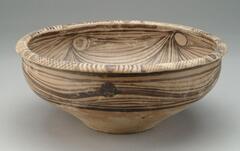 A wide round bowl with an articulated, everted wide rim and a conical lower body on a flat base. It is decorated with wavy linear patterns and black solid dots on the upper half of the exterior, swirling lines with dots on the interior, and cross-hatching on the rim.  