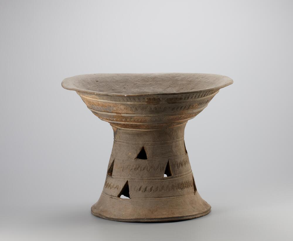 It has wide flared bowl supported by a little flared pedestal foot. Three raised band lines encircle the middle of the bowl. The foot which is separated into 3 parts and has a lot of triangular holes gives the whole bowl stability. There is a tiny wave design on the surface of the bowl and foot<br />
<br />
This is a gray, bowl-shaped, high-fired stoneware vessel stand. The bowl-shaped body flares widely until it spreads horizontally to reach the round rim with grooves. The body is divided by three raised bands into four sections, each of which features a wave design rendered using a multi-tooth comb. The pedestal is divided into five sections by horizontal ridges. Each of the three central sections features four triangular perforations; the lower two of these three central sections are also decorated with wave designs between the perforations. The edge of the pedestal base is narrow and grooved. The inner surfaces of the bowl-shaped body and pedestal show traces of a round inner support anvil that was used