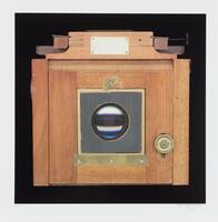A color photograph of an old-fashioned wooden camera, viewed head-on. The lens is black reflective glass and is surrounded by a gray square. A gold knob is to the right of the lens. The background is black.