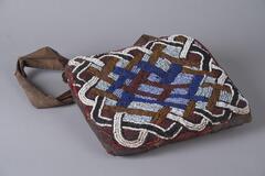 A square bag made of dark red cloth covered with white, blue, black, red, and gold beadwork. The beadwork forms a large interlace pattern. Attached to the top of the bag is a brown cloth handle. 