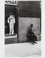 Photograph of a man on a sidewalk leaning against a wall adjacent to a storefront window, which displays a space suit. 