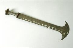 A knife with a wooden handle and a metal blade terminating in a flat top with two points curving outward. Along one side of the blade is a line of white dots and along the other side are remnants of red pigment, possibly a similar line of dots.