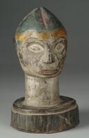 Wooden head with a long neck on a round base. The face is somewhat square and the ears are flat to the sides of the head. The mouth is long and thin, but open, revealing two rows of teeth. The nose is small and the eyes are almond-shaped, inset with metal. The eyebrows are thin and curved. Yellow, blue, and red pigments decorates the forehead and pointed hairstyle. 