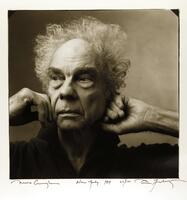 Square black and white photograph showing head, neck and shoulders of a man posed against a blank wall. He is an older man with white curly hair. He is facing toward the viewer, but his eyes gaze to the left as if he is looking at something beyond. His hands rest on either side of his face and his expression is contemplative.