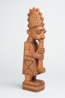 Carved male figure playing a long flute that extends from his mouth past his stomach.&nbsp; The base of the figure has a single-layer chevron pattern.