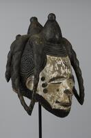 Wooden mask in the form of a female head. The face has traces of white pigment and the lips, nose, eyebrows, and chin are defined with black pigment. There are three small dots in a column on each side of the face. The hair is decorated with spiral designs. Four conical protrusions topped with knobs form the crest of the hairstyle, while four spiral cones curve down from the crest on either side of the head; the two closest to the front on each side are connected at the bottom.