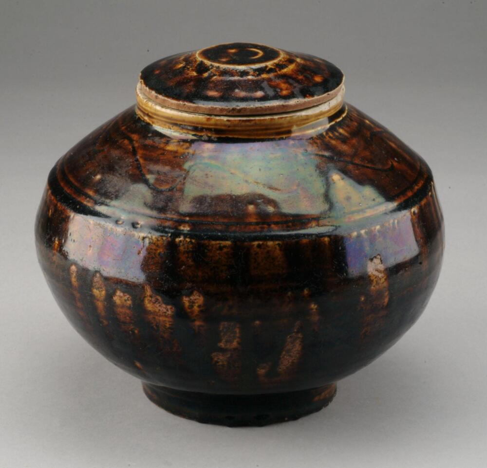 Dark brown glaze has been dripped over the pot from above, allowing it to flow unevenly so that the lighter clay body shows through in an unpredictable pattern. The jar appears rounded in gradations, and has a small circular lid, as well as base.<br />
<br />
This jar is made of white clay and coated with brown-black glaze. No glaze was applied to the inner surface of the lid or the rim of the jar with which the lid comes into contact. The shoulder is incised with a wave design, while an incised line also surrounds the upper part of the body. The glaze is well fused to form a glossy surface. The jar is almost intact.<br />
[Korean Collection, University of Michigan Museum of Art (2014) p.213]