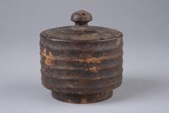 Container made by turning a single piece of wood on a lathe. Lid is crowned with mushroom-shaped knob, and shoulder is formed around mouth to support lid. The lid and body are ribbed. A letter resembling the Chinese character &#39;mok&#39; meaning wood or tree is written in onk on the base of the container. The inside shows traces of having been carved with a knife.<br />
<br />
These containers were made by turning single pieces of wood on a lathe. Their lids are crowned with mushroom-shaped knobs, and shoulder is formed around the mouth to support the lid. Plate 345 (UA2004.60) is decorated with sets of double concentric circles at two places of the upper surface of its lid, and at one place around its side. Also around the body are carved two sets of double circles, and the container is painted with red ocher. Meanwhile, the lid and the body of plate 346 (UA2004.61) are ribbed. A letter resembling the Chinese character &ldquo;mok (木: wood or tree)&rdquo; is written in ink on the base of this container. I