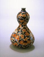 This vase takes the shape of a double gourd, with a large pear-shaped bottom topped by a smaller oval shape. The vase is decorated with overgalze enamels, primarily with an overall pattern of chrysanthemums. The design is also interspersed with plum blossoms, peonies, and auspicious birds.