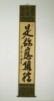 Long calligraphy hanging scroll with green colored backing. Calligraphy paper is faded. Black ink. Stamps in red in the top right corner and near the bottom on the left. A smaller font of calligraphy is set on the left side while the larger set of calligraphy takes up the center of the scroll.