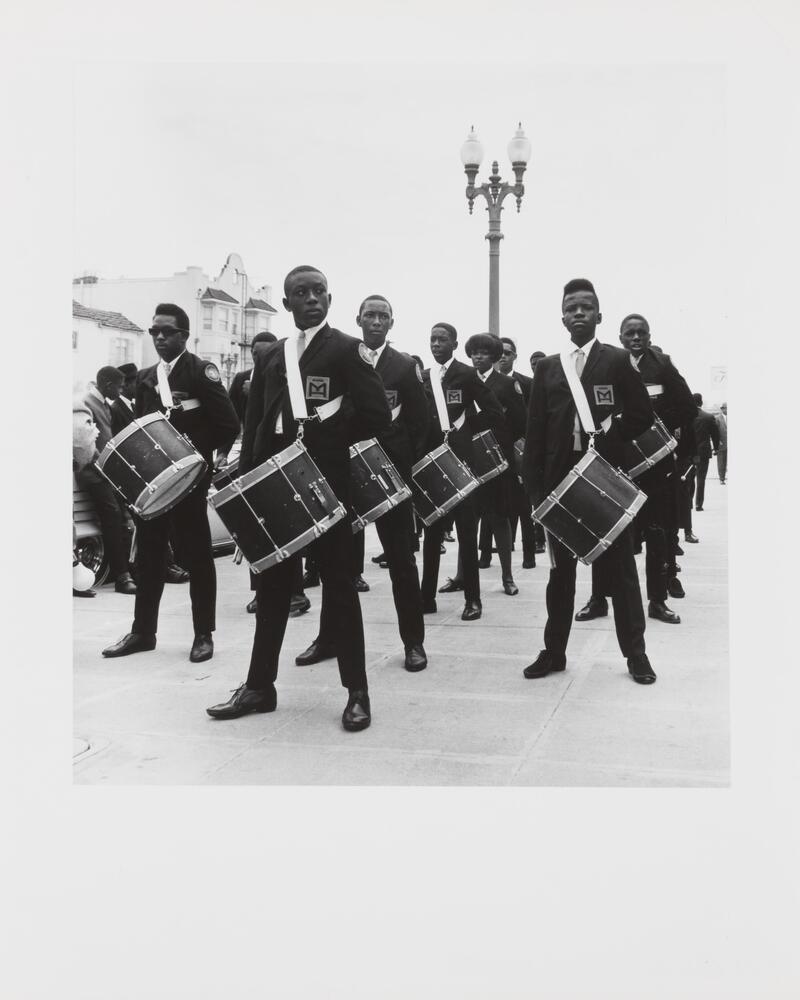 Black and white image of students wearing uniforms wth drums attached by straps. A single lamp post is in the background. 