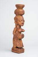 Carved female figure with a child on her back. She carries a basket and balances another container on her head. The base of the piece has a double-layered chevron pattern.&nbsp;