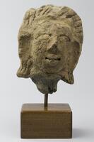 A stone head with full, stylized hair marked by incisions. The ears are elongated and the lips are slightly smiling.