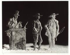 A photograph of three tin figures in a shallow plane, set against a black backdrop. The man on the left stands behind a podium, facing the man in the middle, who is chained. The man on the right holds a whip. 