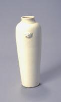 This is a tall, straight-sided, slightly tapered porcelain vase with high narrow shoulders. It has a short neck with an everted rim. Two applique sprig-molded lion heads are placed on opposing sides of the shoulders. The vase is covered in a creamy white glaze. 
