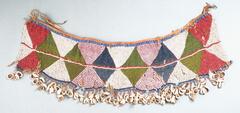 An apron made from multi-colored beadwork, primarily red, white, green, blue, and pink beads in a diamond pattern. The bottom edge of the apron is decorated with a fringe of cowrie shells. 