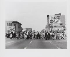 Black and white image of people holding signs while walking across an intersection. 