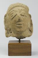 A stone head, smiling slightly, with large eyes. Its right eye droops downward. The ears are elongated and have what appear to be earrings. The top of the head has vertical incisions, indicating styled hair. The verso has a raised column with consumate, inverted v&#39;s.