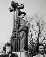 Image of a man standing on the base of a lamp post. He has his hands in his coat pockets and faces to the right of the image. Two women in the lower foreground stare out of the image.