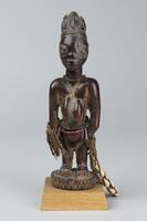 Standing female figure with hands at sides on a round base, which is mounted on a square base. There are strings of beads around the waist, wrists, and neck. Around one wrist is a string of cowrie shells. The pupils of the eyes are metal and the hair is in a tri-lobe shape with patterns of geometric lines. This object has a patina along the creases. 