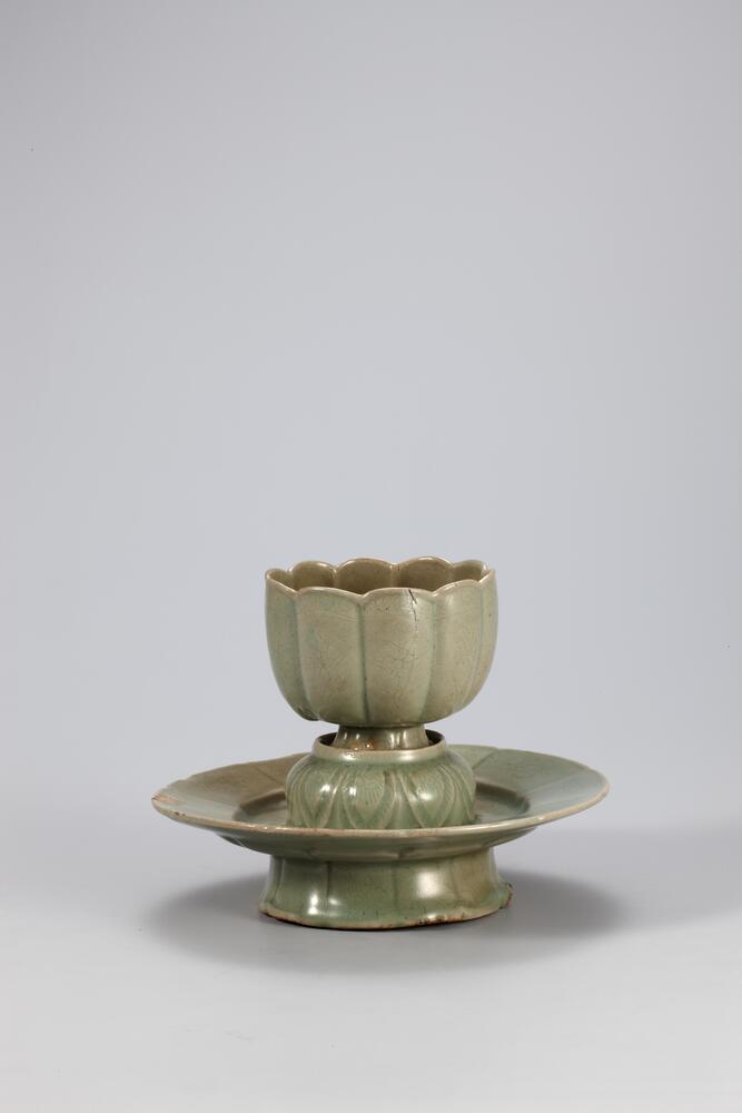 Stoneware lotus-shaped cup and stand with celadon glaze. The cup is shaped in the style of a ten-lobed lotus blossom. On each lobe lies lightly incised chrysanthemum decoration. The cup rests on a pedestal in the design of an inverted lotus flower, which rises from the dish-like base of the stand, mounted on a fluted foot.
<p>This is a flower-shaped cup and stand which offers a good demonstration of the formal splendor of 12th century Goryeo celadon despite yellow-borwn coloration in places. Both the cup and stand have ten lobes and they were produced using molds. On each of the ten lotus petals of the cup and stand is incised a chrysanthemum, and another chrysanthemum design is incised on the upper part of the stand where the cup rests. Around the pedestal on where the cup sits is a band of inverted lotus petals. Each foot of the cup and stand has refractory spur marks.<br />
[<em>Korean Collection, University of Michigan Museum of Art</em> (2014) p.125]<br />
&nbsp;</p>
