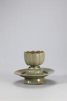 Stoneware lotus-shaped cup and stand with celadon glaze. The cup is shaped in the style of a ten-lobed lotus blossom. On each lobe lies lightly incised chrysanthemum decoration. The cup rests on a pedestal in the design of an inverted lotus flower, which rises from the dish-like base of the stand, mounted on a fluted foot.
<p>This is a flower-shaped cup and stand which offers a good demonstration of the formal splendor of 12th century Goryeo celadon despite yellow-borwn coloration in places. Both the cup and stand have ten lobes and they were produced using molds. On each of the ten lotus petals of the cup and stand is incised a chrysanthemum, and another chrysanthemum design is incised on the upper part of the stand where the cup rests. Around the pedestal on where the cup sits is a band of inverted lotus petals. Each foot of the cup and stand has refractory spur marks.<br />
[<em>Korean Collection, University of Michigan Museum of Art</em> (2014) p.125]<br />
&nbsp;</p>
