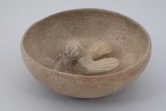 A shallow basin with a flat bottom and gently curved sides, a slightly inverted direct rim, and a central sculpted bird with outstreached wings.  It is covered in a gray-green celadon glaze.