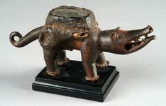 This small figure is that of a dog who has short legs, a thick, curled tail, small ears, and an elongated muzzle. Its mouth is open with pointed teeth bared and tongue slightly hanging. Its eyes include small mirrors and feature the white mineral <em>kaolin</em>. On the dog’s back is a large, mirror-topped medicine pack which has been sealed with resin.  There are two recesses on top of both hind legs which also likely held packets of medicine.  Near the top of the two front legs are hollow projections, which have broken off.