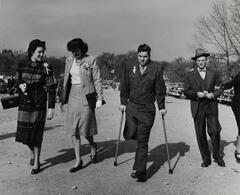 Image of two women and two men walking in a parking lot. One man uses crutches, is missing his right leg, and wears a World War II army uniform.&nbsp;