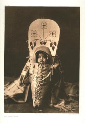 This is a portrait of a baby in a cradleboard, taken against a dark backdrop. The baby is tied into the carrying device, which is decorated with floral motifs. A patterned blanket is draped around the base of the cradleboard.