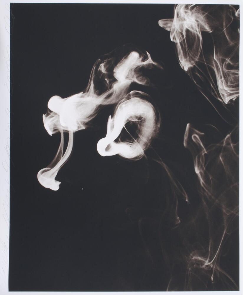 Square black and white photograph depicting wisps of smoke and a smoke ring that float against a black backdrop.