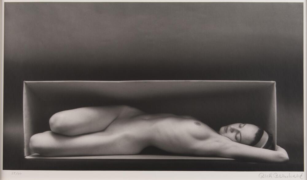 This monochrome photograph of a nude woman shows her reclining, knees bent; her figure framed by a large box. 