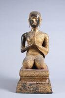 Kneeling statue of Buddhist monk devotee Mogallana or Shariputra on a pedestal with round face, hair in tight curls, elongated ears, and hands placed together in mediatation.  The pedestal is decorated with lotus petals, and the devotee wears an intricate, close-fitting robe.
