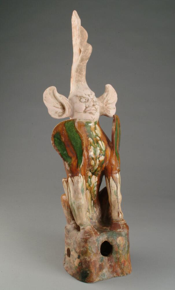 An earthenware figure of an anthropomorphic form consisting of a lion-like body with strong hoofed feet, wings, and a human face with central flame-like horns and large, furry and pointed ears. It is seated on a rock-like base, and except for the head it is covered in amber, green, and cream runny glazes. One of a pair with 2004/2.132.1.